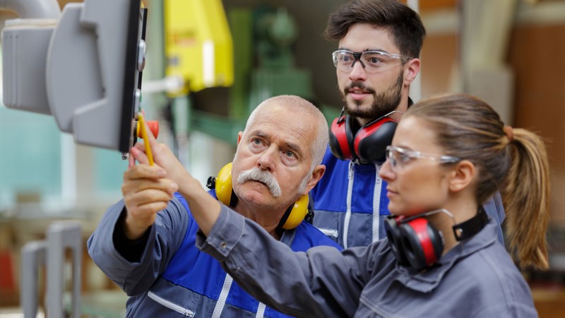 Picture of a scene in a workshop in which two men and a woman are looking at a device being operated by the woman and the older man. All three are wearing work clothes and protective equipment.