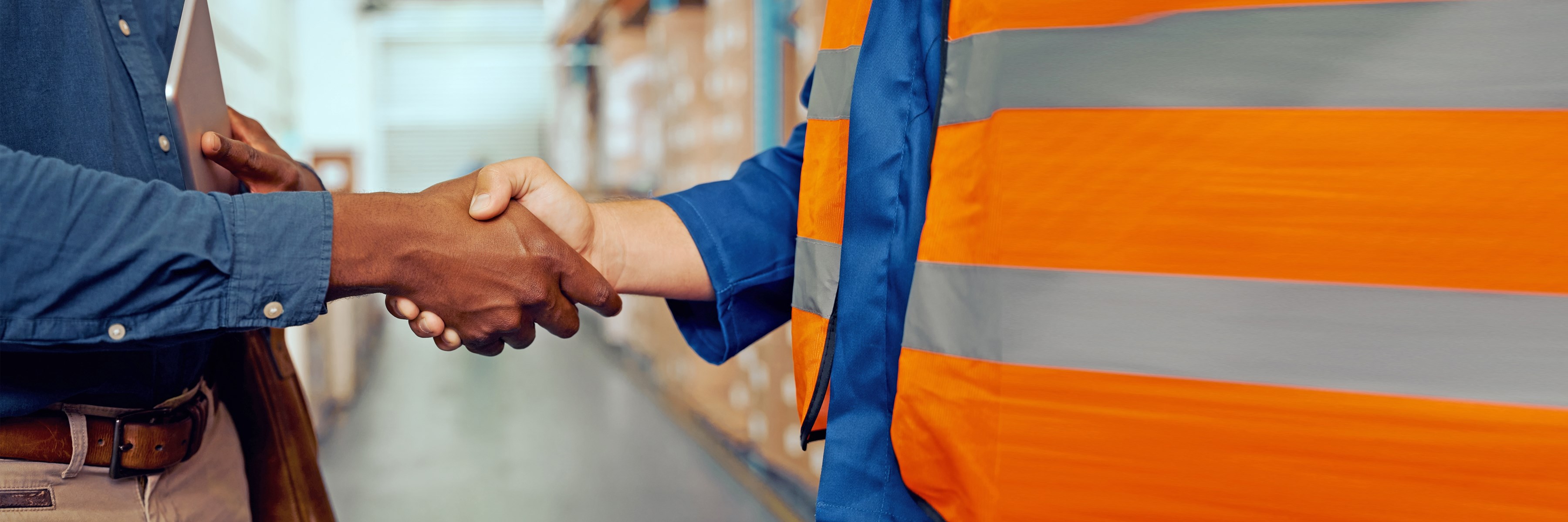 Two people shake hands as partners. The picture shows a close-up of only the two hands. The person on the left is wearing a blue shirt and the person on the right is wearing an orange safety vest. The surroundings of a workshop can be seen in the background.)